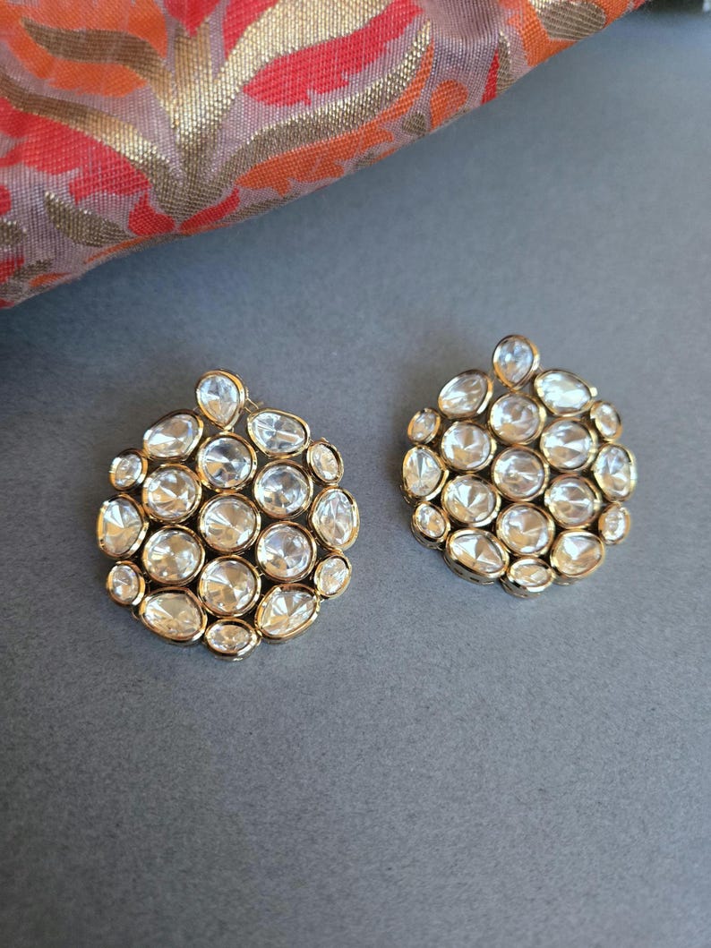 May include: A pair of gold-coloured stud earrings. Each earring has a cluster of clear, faceted stones in various shapes, set in a circular arrangement. The earrings are displayed on a grey surface, with patterned fabric in the background.