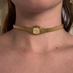 Puede incluir: Un reloj dorado usado como gargantilla, con una correa de metal texturizado y una pequeña esfera rectangular con números y manecillas negras. La caja del reloj está integrada en el centro de la correa metálica delgada y flexible.