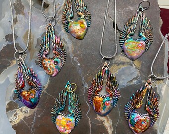 Könnte beinhalten: Eine Sammlung herzförmiger Anhänger mit schillernden Flügeln, jeweils an einer silbernen Kette. Die Herzen zeigen eine Reihe von Farben, darunter Gold, Blau und Lila. Die Anhänger werden auf einer strukturierten, braunen Oberfläche präsentiert.