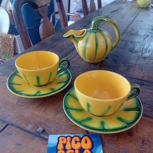 Op de afbeelding: Een gele keramische theeset met groene accenten. De set bevat twee kopjes en schotels, en een kleine kan. De kopjes en kan hebben een geribbeld ontwerp. Een sticker met de tekst "PICO BELO" ligt op de tafel.