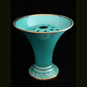 Può includere: Un vaso di fiori in ceramica turchese con la parte superiore svasata e una base rotonda. Il vaso ha un bordo dorato e dettagli dorati a forma di onda. La parte superiore ha più fori per disporre i fiori. Il vaso è su sfondo nero.
