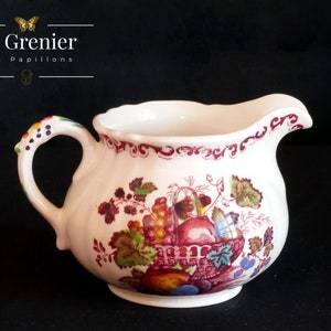 Puede incluir: Una jarra de crema de cerámica blanca con un diseño floral rojo y verde que presenta una cesta de frutas. La jarra tiene un borde festoneado y un asa con un diseño floral.