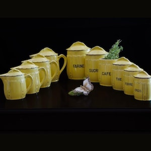 Peut inclure: Ensemble de pots et de pichets de cuisine en céramique jaune avec des bordures noires. Les pots sont étiquetés avec les mots "FARINE", "SUCR", "CAFE", "THE", "POIVRE" et "EPICES". Un papillon décoratif est posé devant.