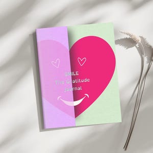 Può includere: Un diario della gratitudine con un design a cuore rosa e la parola "SMILE" stampata sulla copertina. Il diario ha un dorso lilla chiaro e una copertina posteriore verde chiaro. Sono presenti anche un piccolo cuore e una bocca sorridente.