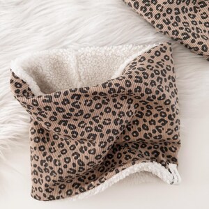 Könnte beinhalten: Ein beigefarbener und schwarzer Schal mit Leopardenmuster und einem weichen, weißen Innenfutter. Der Schal aus Rippstoff ist für Wärme konzipiert. Das Design zeigt ein sich wiederholendes Muster aus schwarzen Flecken. Zum Tragen um den Hals.