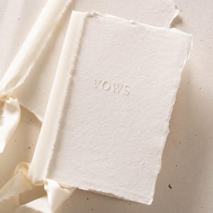 Puede incluir: Un libreto de color crema con bordes irregulares y una superficie texturizada. La palabra VOWS está grabada en relieve en el centro de la portada. Una cinta a juego de color crema está atada alrededor del lomo, con bordes deshilachados.