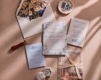 May include: Wedding invitation suite featuring a floral-lined envelope, invitation cards with blue floral accents, a reply card, and an information card. A wax seal, ring box, and vintage stamps complete the arrangement. The suite is arranged on a textured, pink surface.