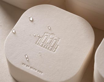 Puede incluir: Una pila de posavasos cuadrados blancos con bordes redondeados. El posavasos superior tiene un pequeño diseño en relieve de un edificio y el texto "To be with you".