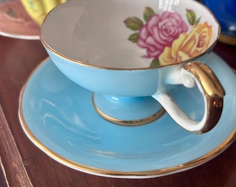 Puede incluir: Juego de taza y platillo azul claro con ribete dorado. La taza presenta un diseño floral con rosas rosas y amarillas. El asa y el borde son dorados.