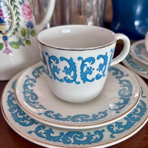 Könnte beinhalten: Weißes Tee-Service mit blauem Rankenmuster und goldenem Rand. Die Tasse steht auf zwei passenden Untertassen. Eine weiße Teekanne mit floralen Akzenten ist im Hintergrund zu sehen.
