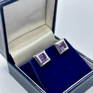 May include: A pair of square-cut purple amethyst stud earrings set in polished silver-toned metal. The earrings are displayed inside a dark blue hinged jewellery box with a white satin interior lining and a deep blue velvet base insert.
