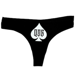 Könnte beinhalten: Schwarzer Tanga mit einer weißen Pik-Grafik auf der Vorderseite. Das Pik-Symbol enthält die Buchstaben QOS, die in einer fetten, schwarzen Schrift im gotischen Stil gedruckt sind. Das Kleidungsstück hat einen breiten Bund und einen klassischen Tanga-Schnitt.