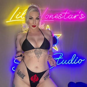 May include: A black bikini set featuring a triangle top and a high-cut bottom. The bottom piece displays a red spade symbol with devil horns on the front. The neon sign in the background reads Lil Lonestar's Studio in yellow, pink, and blue.