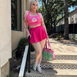 May include: A bright pink halter crop top featuring a white spade graphic with wings, paired with a matching pleated mini skirt. The outfit is accessorized with white socks, silver sneakers, and a green and pink patterned tote bag with pink straps.