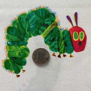 Puede incluir: Un diseño de tela con una colorida ilustración de una oruga. La oruga es principalmente verde con acentos rojos, amarillos y morados. Una moneda estadounidense está al lado de la oruga como referencia de tamaño.