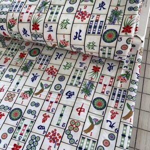 May include: White fabric with a repeating pattern of Mahjong tiles. The tiles feature Chinese characters, bamboo, flowers, and circles in red, green, and blue. The fabric is rolled up.