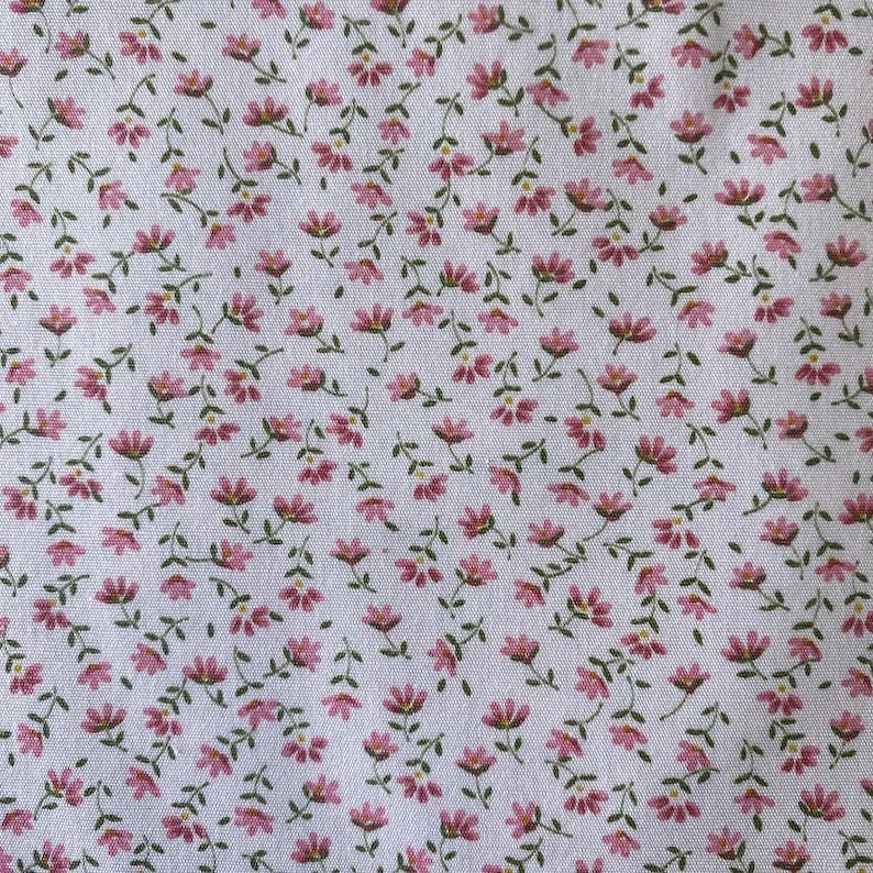 May include: White fabric with a repeating pattern of small pink flowers and green stems. The floral design is evenly distributed across the surface, creating a delicate and feminine aesthetic. The fabric appears to be cotton.