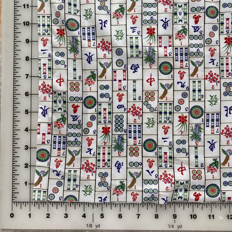 May include: White fabric with a repeating pattern of Mahjong tiles. The tiles feature various symbols, numbers, and illustrations in red, green, blue, and black. The fabric is measured with a ruler in inches.