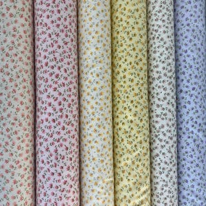May include: A collection of fabric rolls featuring floral patterns in various colors. The fabrics display small, repeating flower designs in shades of pink, yellow, and blue on a white background. The rolls are tightly wound and vertically arranged.