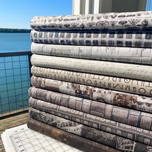 May include: A stack of fabric rolls with various vintage-style patterns. The designs include butterflies, letters, and ticket stubs in shades of brown, cream, and gray. The fabrics are neatly stacked, showcasing the different patterns.