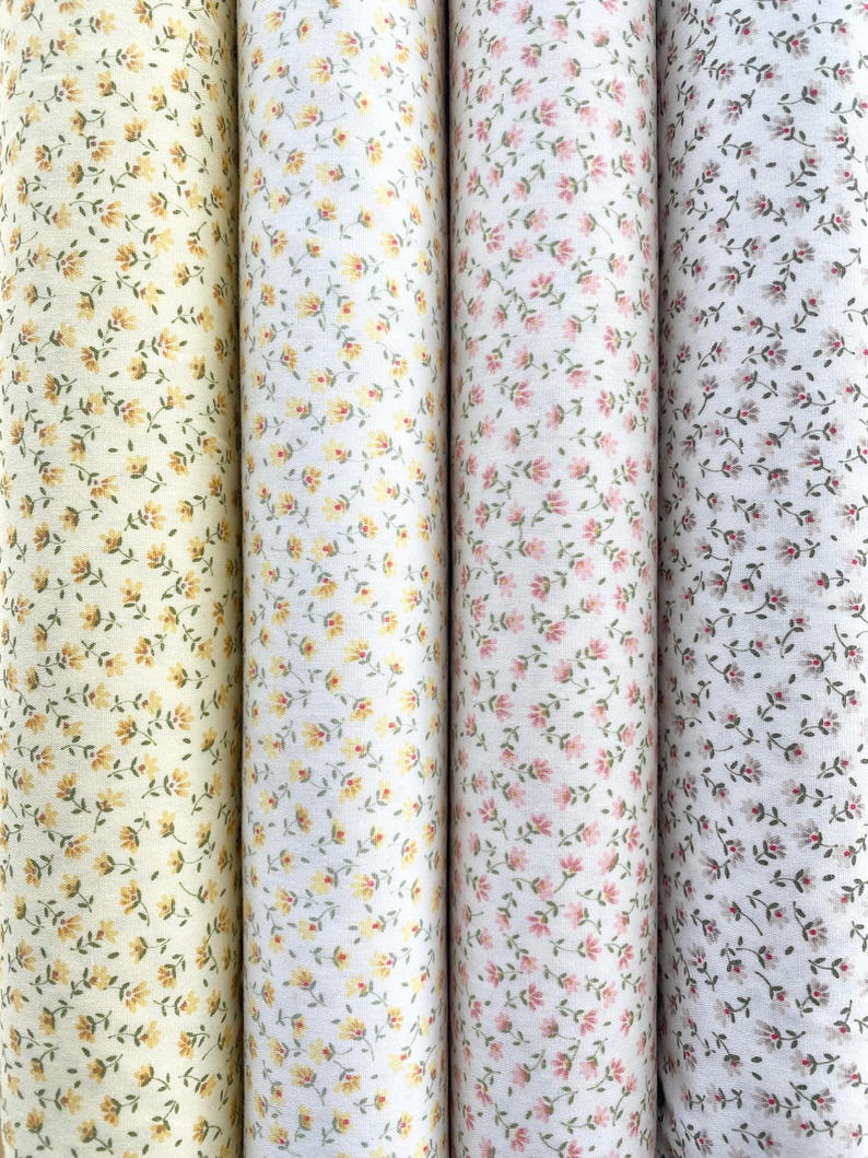 May include: Four rolls of fabric with floral patterns. The rolls display a variety of colors, including yellow, white, and pink, each adorned with small, delicate flower designs. The fabric is tightly rolled, showcasing the repeating floral patterns.
