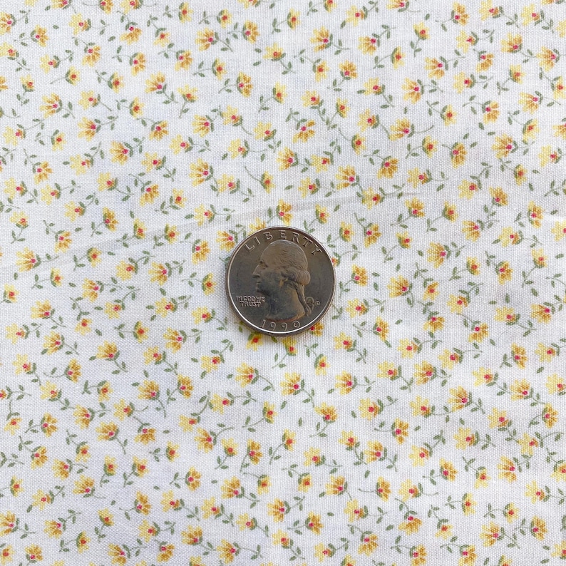 May include: Close-up of white fabric with a repeating pattern of small yellow flowers and green stems. A U.S. quarter coin is placed on the fabric, showing the year 1990. The fabric has a vintage, floral design.