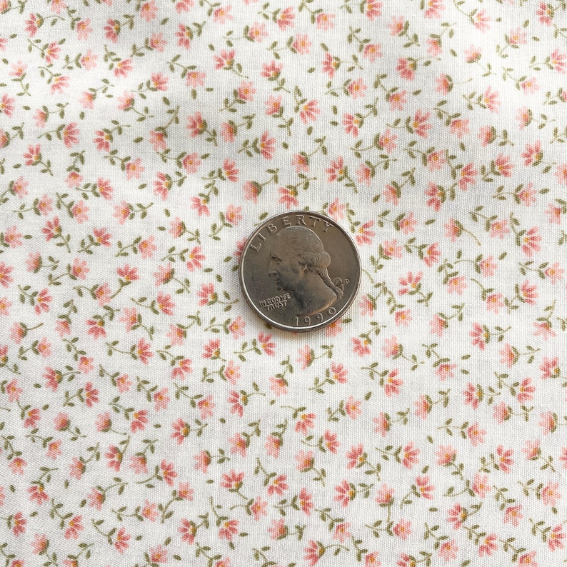 May include: Close-up of a white fabric with a repeating pattern of small pink flowers and green stems. A U.S. quarter coin is placed on the fabric for scale. The floral design is delicate and detailed, creating a vintage or cottagecore aesthetic.