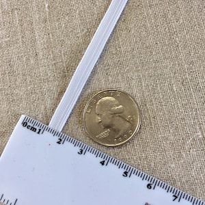 May include: A close-up of white elastic band next to a U.S. quarter coin and a ruler. The elastic band is approximately 0.25 inches wide. The ruler shows measurements in centimeters. The quarter coin is silver in color.