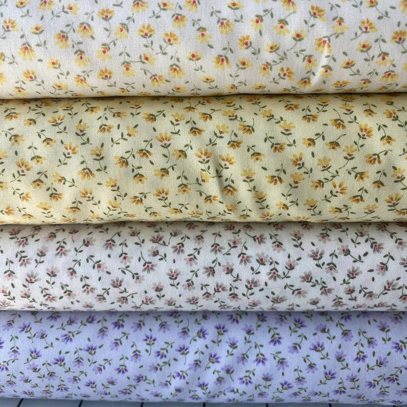 May include: Four rolls of floral fabric in various colors. The top roll is cream-colored with yellow and red flowers. The second roll is yellow with yellow flowers. The third roll is white with pink flowers. The bottom roll is lavender with purple flowers.