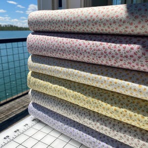 May include: A stack of fabric rolls in various colors, each covered in a small floral pattern. The colors include white, pink, yellow, and lavender. The fabric is likely cotton or a similar material, suitable for quilting or sewing projects.