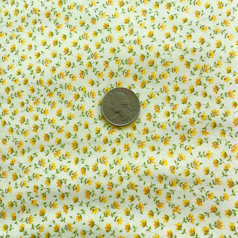 May include: Close-up of a fabric with a repeating pattern of small yellow flowers and green leaves on a cream background. A U.S. quarter coin is placed on the fabric for size reference.
