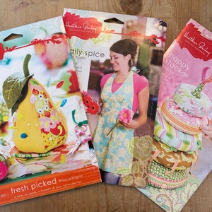 May include: Three sewing patterns featuring pincushions and an apron. The first pattern is for a pear, apple, tomato, and strawberry pincushion set. The second pattern is for a floral apron called "Daily Spice". The third pattern is for a stack of ring toys called "Happy Stacker".