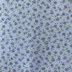 May include: Close-up of a fabric with a repeating floral pattern. Small purple flowers with yellow centers and green stems are scattered across a light blue background. The fabric has a delicate, vintage aesthetic.