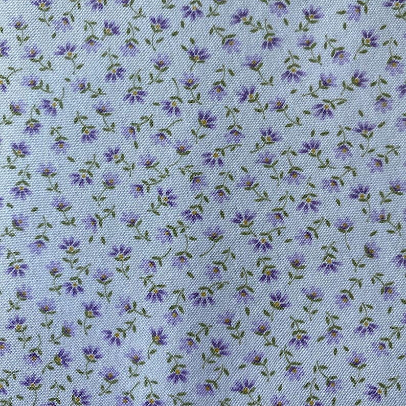 May include: Close-up of a fabric with a repeating floral pattern. Small purple flowers with yellow centers and green stems are scattered across a light blue background. The fabric has a delicate, vintage aesthetic.
