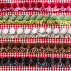 Könnte beinhalten: Fünf Reihen verschiedener farbiger Borten mit Bommeln. Die Borte befindet sich auf einem rot-weiß karierten Hintergrund. Die Farben der Borte sind rosa, grün, weiß, braun und schwarz.