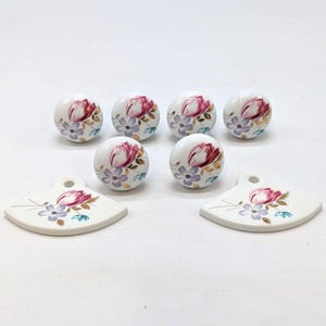May include: A set of six round ceramic cabinet knobs and two fan-shaped pulls. Each piece features a white base with a floral design of pink tulips, purple flowers, and green leaves. The knobs and pulls are arranged on a white surface.