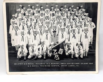 May include: Black and white photograph of a group of U.S. Navy sailors in uniform, posing for a group photo. The photo includes a flag with the text "A107" and the anchor symbol. The photo's text reads "CO. A107 U.S. NAVAL RESERVE R.E. MURPHY, BMGC.USN.CO.CMDR. 24 JUNE 1952. U.S. NAVAL TRAINING CENTER, GREAT LAKES, ILL."
