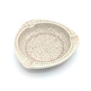 May include: A speckled, off-white ceramic ashtray with a triangular shape and three cigarette rests. The ashtray has a textured rim and a speckled interior with brown flecks. The design is simple and functional.