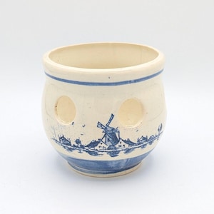 May include: A cream-colored ceramic pot featuring a blue transferware pattern of a Dutch landscape with a windmill. The pot has a rounded body, a wide opening, and three circular cutouts spaced evenly around the upper portion of the exterior.
