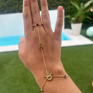 Peut inclure: Ensemble de bijoux de main doré avec une bague et un bracelet reliés par une chaîne délicate. La bague et le bracelet comportent des accents de pierres précieuses bleues. Le bracelet comprend un charm en forme de main de Hamsa.