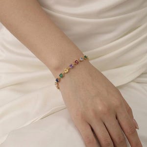 Peut inclure: Un bracelet à chaîne doré orné d'une série de pierres précieuses multicolores de taille carrée, notamment des pierres bleues, vertes, jaunes, violettes et rouges, reliées entre elles dans un motif répétitif qui entoure le poignet avec éclat.