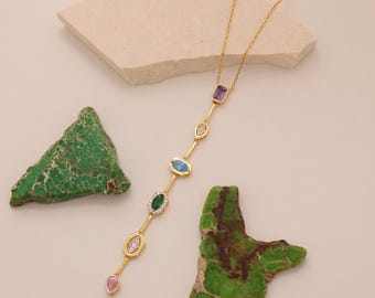 May include: A gold-colored necklace with a vertical bar design, featuring various colorful gemstones. The gemstones include purple, clear, blue, green, and pink. The necklace is displayed on a neutral background with green and beige decorative elements.