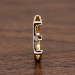 Peut inclure: Une bague dorée en forme de lettre "E", avec un petit diamant. La bague a un design bicolore, avec un contour argenté. La bague est présentée sur une surface en bois.