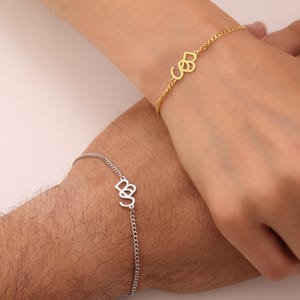 May include: Two personalised bracelets, one gold and one silver, each featuring intertwined initials. The gold bracelet is on a delicate chain, while the silver bracelet has a similar chain design. The bracelets are worn on wrists, showcasing a custom jewellery design.