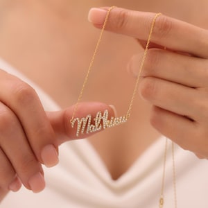 Può includere: Una collana color oro con il nome "Matthew" scritto in corsivo, incastonata con piccole pietre scintillanti. La collana è tenuta da una persona, mettendo in risalto la delicata catena e il ciondolo. Un gioiello personalizzato.