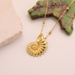 May include: A gold-colored necklace featuring a nautilus shell pendant. The pendant has detailed ridges and a spiral design. The necklace chain is delicate, with small ball accents. The background includes a green and brown stone and a white surface.