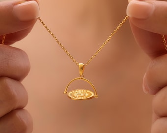 May include: A gold-colored pendant necklace featuring a circular charm with a sunburst design. The charm is suspended from a delicate chain and held by a person's hands. The necklace is a jewelry item.