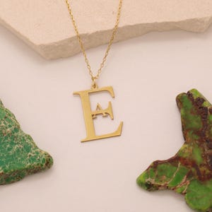May include: A gold-colored necklace featuring a large letter "E" pendant. The pendant is suspended from a delicate chain. The necklace is displayed on a light-colored surface, with green and brown decorative elements on either side.