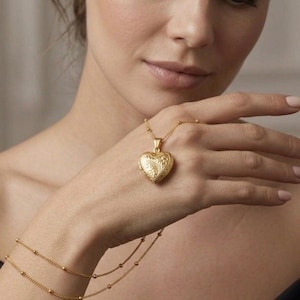May include: A gold heart-shaped locket necklace with an intricate floral pattern. The locket is held in a hand, with a fine gold bracelet on the wrist. The jewellery is set against a soft, neutral background.