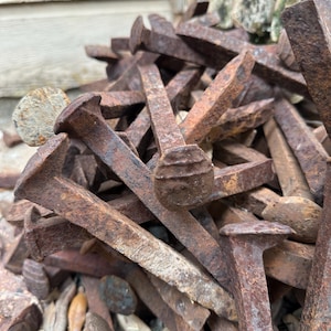 May include: A pile of weathered, rusty railroad spikes. The spikes have large, flat heads and tapered bodies, showing signs of age and exposure. The color is a mix of brown and orange, with some areas of darker rust.